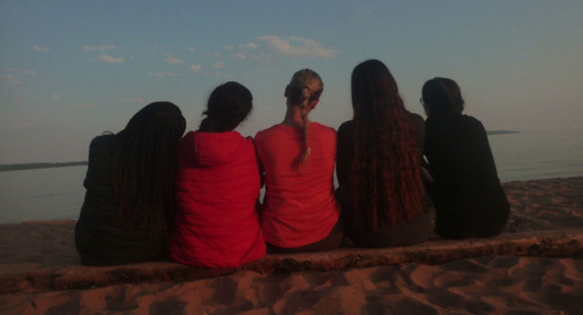 Five people sit close together with their backs facing the camera. They are looking out over a body of water. 